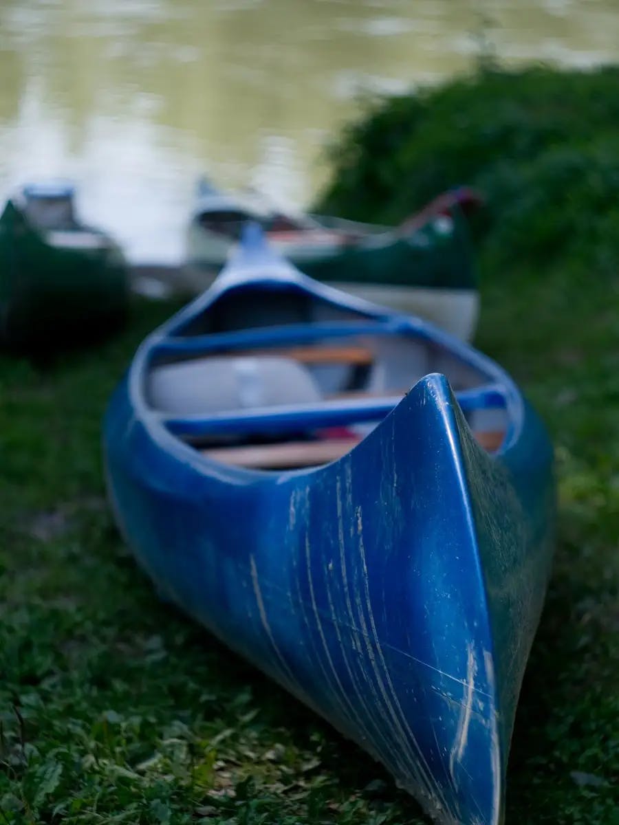 Blue canoe resting on grass near a lake, with other canoes in the background.