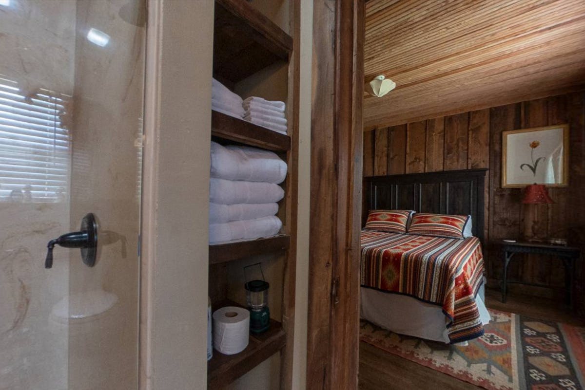View from a bathroom with shelves of towels into a cozy bedroom with a striped bedspread and wooden walls.