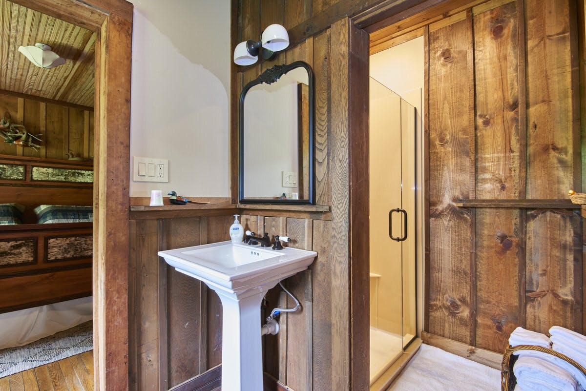Rustic bathroom with a pedestal sink, mirror, and view into a bedroom with wooden walls.