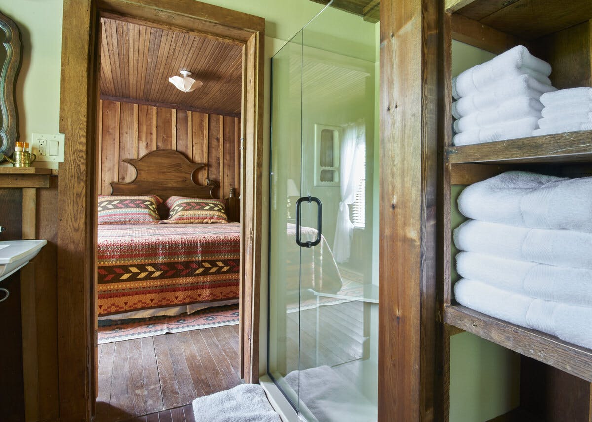 View from a bathroom with shelves of towels into a rustic bedroom with a colorful patterned bedspread and wooden walls.