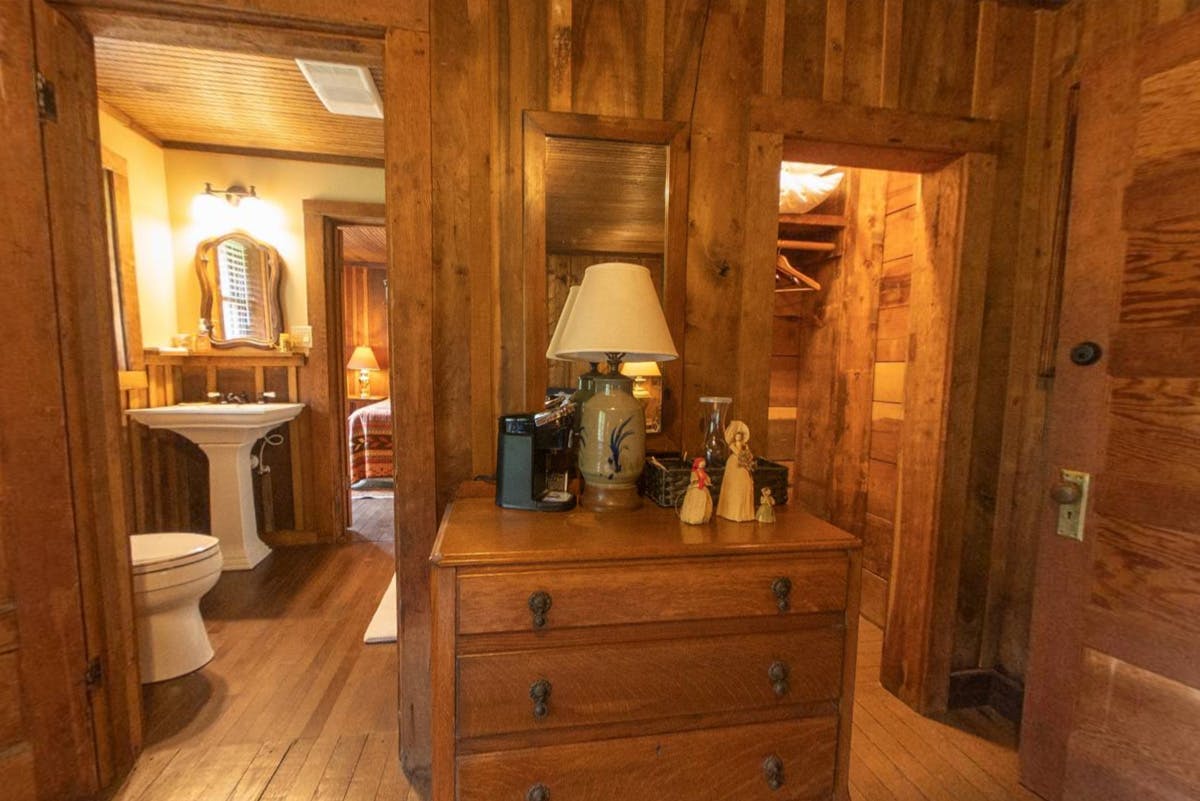 Rustic hallway with a dresser, lamp, and view into a bathroom with a pedestal sink and a bedroom with wooden walls.