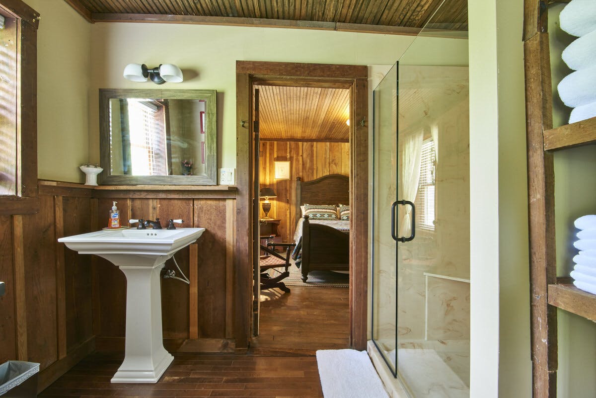 Rustic bathroom with a pedestal sink, glass shower, and view into a bedroom with a wooden headboard.