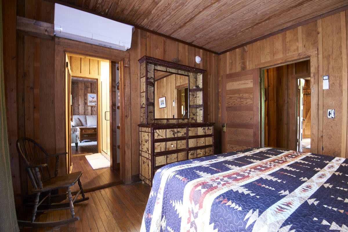 Rustic bedroom with a patterned bedspread, wooden dresser with a mirror, and wooden walls.