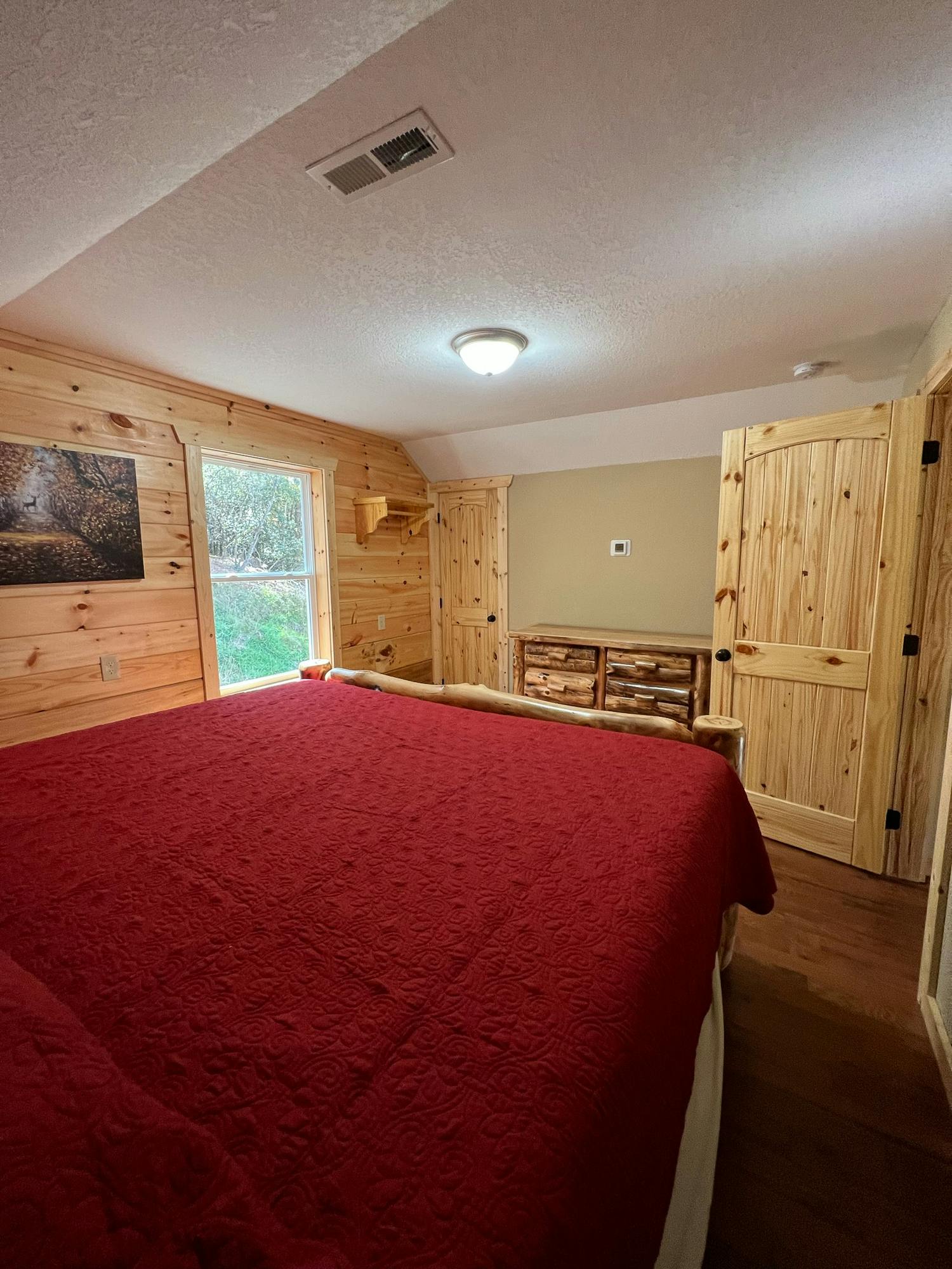 Cozy bedroom with wooden walls, a red bedspread, and natural light from a window.